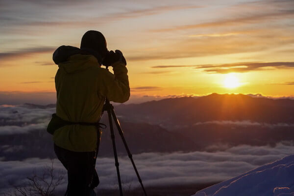 三脚を構えて朝日を撮影する登山者